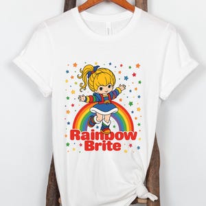 May include: White t-shirt featuring a cartoon illustration of Rainbow Brite. The design includes a character standing on a rainbow with colorful stars. The text "Rainbow Brite" is below the character.