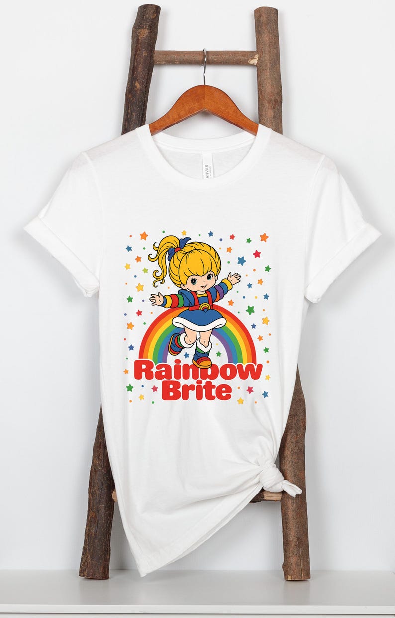 May include: White t-shirt featuring a cartoon illustration of Rainbow Brite. The design includes a character standing on a rainbow with colorful stars. The text "Rainbow Brite" is below the character.