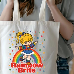 May include: White tote bag featuring a cartoon illustration of Rainbow Brite standing on a rainbow. The design includes colorful stars and the text "Rainbow Brite" in red. The bag has two white handles.
