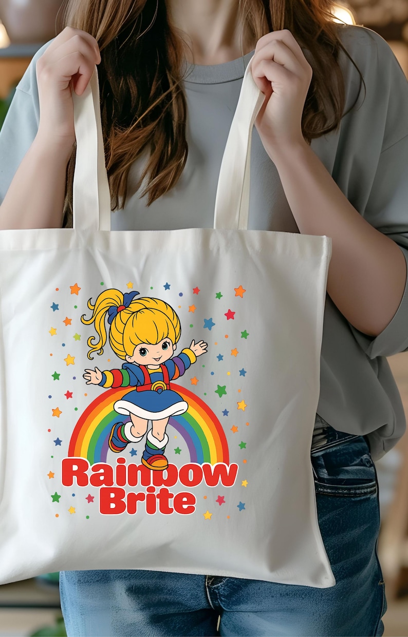 May include: White tote bag featuring a cartoon illustration of Rainbow Brite standing on a rainbow. The design includes colorful stars and the text "Rainbow Brite" in red. The bag has two white handles.