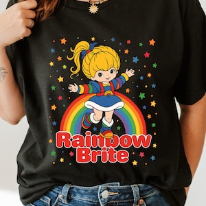 May include: Black t-shirt featuring a cartoon image of Rainbow Brite standing on a rainbow with colorful stars. The text "Rainbow Brite" is displayed in red and white. The character has blonde hair and is wearing a blue dress.