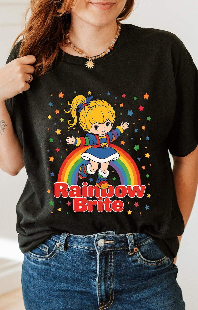 May include: Black t-shirt featuring a cartoon image of Rainbow Brite standing on a rainbow with colorful stars. The text "Rainbow Brite" is displayed in red and white. The character has blonde hair and is wearing a blue dress.