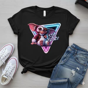 May include: Black t-shirt featuring a cartoon Gremlin character with the text "Stay Fuzzy" inside a neon triangle. The design includes pink, brown, and white colors. The shirt is paired with blue jeans and white sneakers.