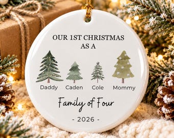 May include: A round white ceramic ornament featuring the text OUR 1ST CHRISTMAS AS A FAMILY OF FOUR - 2026. It displays four watercolor-style evergreen trees labeled Daddy, Caden, Cole, and Mommy, suspended by a thin gold string for hanging.
