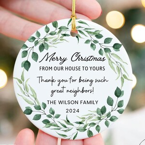May include: White ceramic Christmas ornament with a green leafy wreath design. The ornament reads "Merry Christmas from our house to yours. Thank you for being such great neighbors! The Wilson Family 2024".