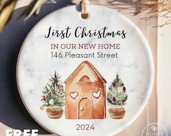 Peut inclure: Une décoration ronde en céramique blanche avec une texture marbrée, illustrée à l'aquarelle d'une maison en pain d'épices entourée de deux sapins de Noël décorés. Le texte indique First Christmas IN OUR NEW HOME 146 Pleasant Street 2024.