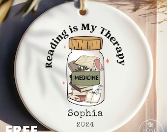 May include: A round white ceramic ornament featuring a graphic of a glass jar filled with books and a label reading MEDICINE. The text Reading is My Therapy arches above the jar, with the name Sophia and the year 2024 printed below it.