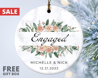 May include: A round white marble-patterned ceramic ornament featuring a floral arrangement of pink roses and green pine branches. The text "Engaged" is printed in black script, followed by the names "MICHELLE & NICK" and the date "12.21.2022".