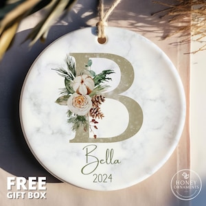 May include: A round white ceramic ornament with a marble pattern, featuring a large gold letter B decorated with a floral arrangement of a white pumpkin, a rose, pine cones, and greenery. The name Bella and the year 2024 are printed below the letter.