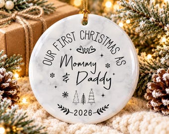 May include: A round white marble-patterned ceramic ornament with black text reading Our First Christmas As Mommy & Daddy 2026, featuring illustrations of three small pine trees, snowflakes, stars, and hearts, suspended by a gold string.
