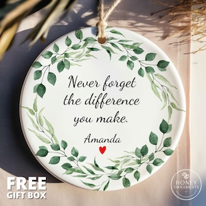 May include: A round white ceramic ornament featuring a green watercolor leaf wreath design. The text reads, Never forget the difference you make. Amanda, with a small red heart below the name. A jute twine loop is attached at the top for hanging.