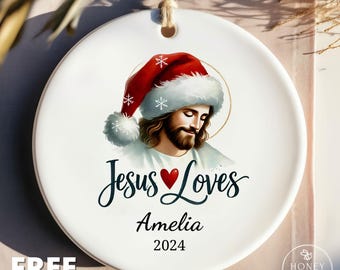 May include: A round white ceramic ornament featuring an illustration of a bearded figure wearing a red and white Santa hat with snowflakes. Below the image, the text reads Jesus Loves, followed by the name Amelia and the year 2024.
