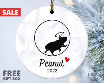 May include: A round white ceramic ornament with a gray marble pattern. The center features a black silhouette of a guinea pig with a halo inside a circle. Below the graphic, the name Peanut is printed in black with a small red heart, followed by 2023.