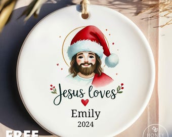 May include: A round white ceramic ornament featuring a cartoon illustration of a bearded figure wearing a red Santa hat with a gold halo. Below the figure, the text reads Jesus loves, followed by the name Emily and the year 2024 in black font.