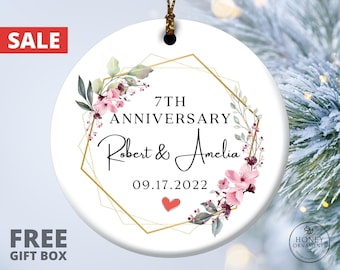 May include: A round white ceramic ornament featuring a gold geometric frame, pink floral accents, and black text reading 7TH ANNIVERSARY, Robert & Amelia, and 09.17.2022, with a small red heart icon at the bottom and a gold hanging string.