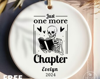 May include: A round white ceramic ornament featuring a black graphic of a skeleton reading a book. The text reads Just one more Chapter, Evelyn, and 2024. The ornament has a hole at the top with a jute twine string for hanging, and black heart accents.