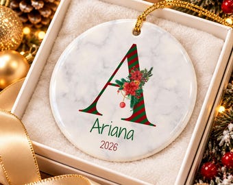 May include: A round white marble-patterned ceramic ornament featuring a large letter A with red and green diagonal stripes. The letter is decorated with red poinsettias and pine branches. The name Ariana and the year 2026 are printed below.