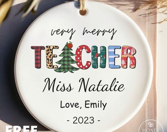 May include: A round white ceramic ornament with a jute string hanger. The text reads very merry TEACHER with the letter A replaced by a green Christmas tree. Below is the text Miss Natalie, Love, Emily, and the year 2023 in black font.
