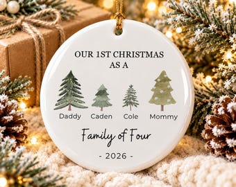 May include: A round white ceramic Christmas ornament featuring the text OUR 1ST CHRISTMAS AS A, followed by four illustrated evergreen trees labeled Daddy, Caden, Cole, and Mommy, with the text Family of Four and the year 2026 below.