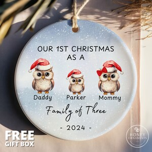 May include: A white ceramic Christmas ornament with a blue background and a snowy design. It features three cartoon owls wearing Santa hats and a red bow. The text reads "Our 1st Christmas as a Family of Three - 2024 - "