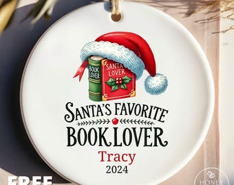 May include: A round white ceramic ornament featuring a red Santa hat resting on a green and red book labelled Santa Lover and Book Lover. Below the book, the text reads Santa's Favorite Book Lover, followed by the name Tracy and the year 2024.