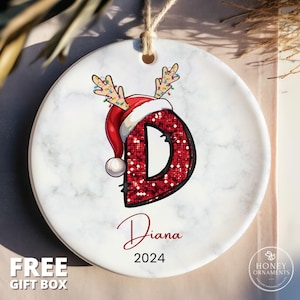 May include: A white ceramic Christmas ornament with a red sparkly letter D, a red Santa hat with white trim, and reindeer antlers with colorful lights. The ornament is personalized with the name "Diana" and the year "2024".