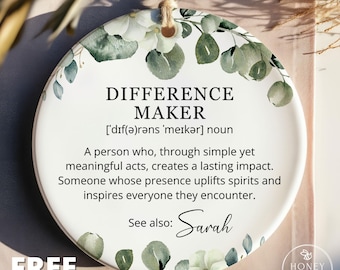 May include: A round white ceramic ornament featuring a watercolor eucalyptus leaf and white flower border. The text reads: DIFFERENCE MAKER, a definition of the noun, and the phrase See also: Sarah. A jute twine loop is attached at the top.