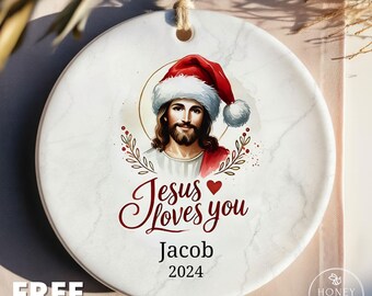 May include: A round white ceramic ornament with a marble texture featuring an illustration of Jesus wearing a Santa hat. Below the image, the text reads Jesus loves you, followed by the name Jacob and the year 2024 in black serif font.