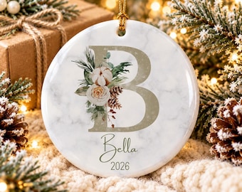 May include: A round white ceramic ornament with a marble pattern featuring a large gold letter B, a floral arrangement with pine cones and greenery, the name Bella in script, and the year 2026, suspended by a gold string.