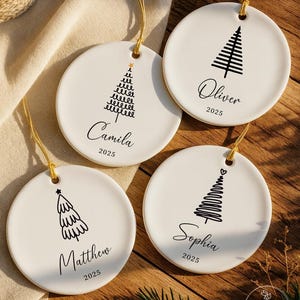 May include: Four round white ceramic ornaments with gold hanging strings. Each features a black line-art Christmas tree, a name in script font, and the year 2025. The names are Camila, Oliver, Matthew, and Sophia. One tree has a gold star.