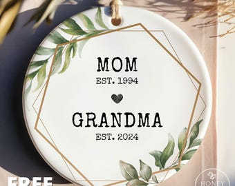 May include: A round white ceramic ornament featuring a gold geometric frame, green watercolor leaves, and black text reading MOM EST. 1994, a small heart, and GRANDMA EST. 2024. The ornament hangs from a rustic twine loop at the top center.