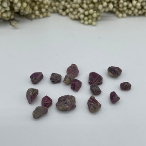 Könnte beinhalten: Eine Sammlung kleiner, rauer, rosa und violetter Rubin-Edelsteine. Die Steine sind auf einer weißen Oberfläche verstreut.