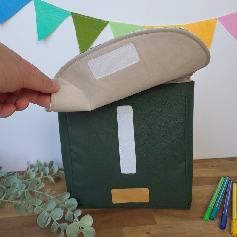 May include: A green lunch bag with a white exclamation point and a brown leather patch. The bag is open and the inside is beige.