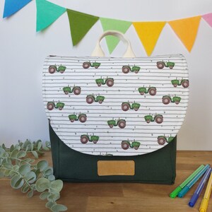May include: A green and white children's backpack with a tractor pattern. The backpack has a white flap with a green tractor pattern and a dark green bottom. The backpack has a cream-colored handle.