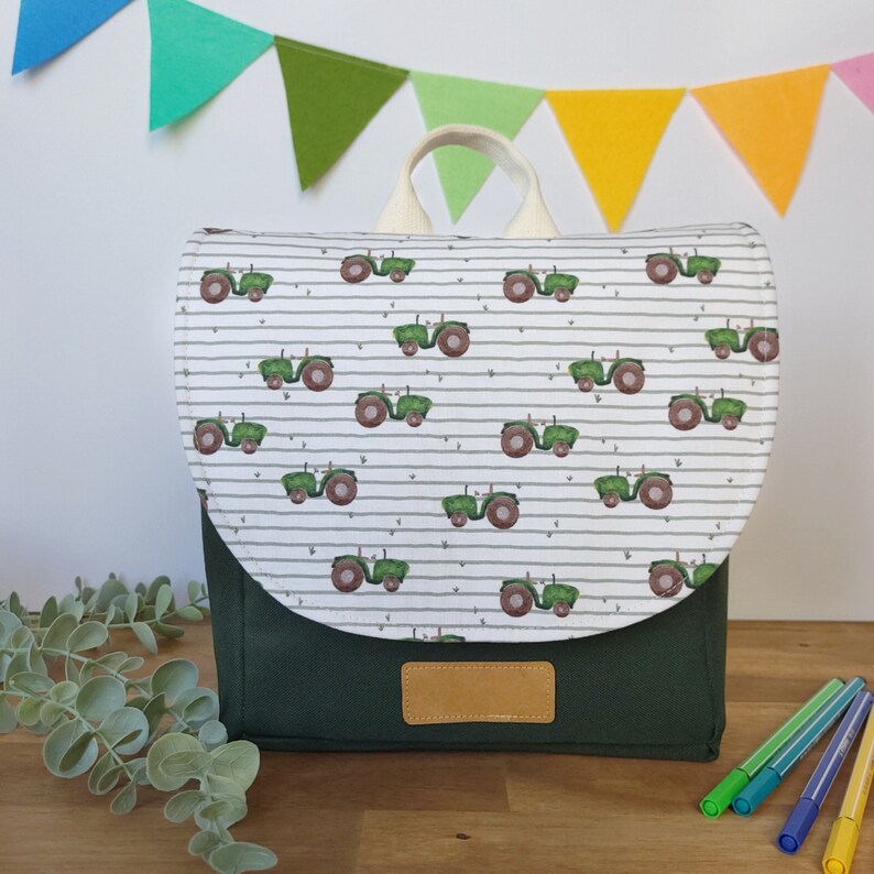 May include: A green and white children's backpack with a tractor pattern. The backpack has a white flap with a green tractor pattern and a dark green bottom. The backpack has a cream-colored handle.