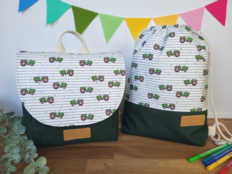 May include: A green and white backpack and a green and white drawstring bag with a tractor pattern. Both bags have a green bottom and a brown leather label.