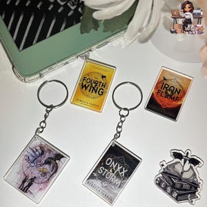 May include: A collection of book-themed keychains and a sticker. The keychains feature book covers with titles like "Fourth Wing," "Iron Flame," and "Onxx Storm." The sticker shows a stack of books with a bat on top. The keychains have silver chains and rings.