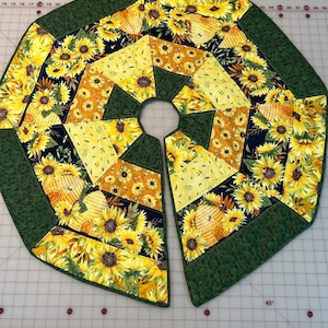 Op de afbeelding: Een gesplitste, zeshoekige boomrok met een zonnebloem- en honingraatpatroon. De rok is voornamelijk geel en zwart, met een groene rand. Het ontwerp bevat gedetailleerde afbeeldingen van zonnebloemen en honingraten.