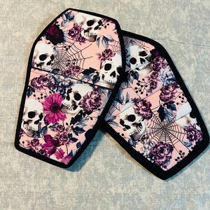 Puede incluir: Dos agarraderas de olla en forma de ataúd de color rosa y negro con un patrón floral y de calaveras. El patrón incluye rosas rosas, flores moradas y calaveras blancas con ojos negros.