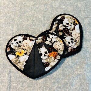 Puede incluir: Dos agarraderas de olla de corazón negro con un estampado de calavera y flores. Las agarraderas están hechas de tela y tienen un borde negro.