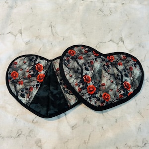 May include: Two heart-shaped pot holders with a black trim. The fabric features a gray background with a pattern of red roses and bare tree branches. The pot holders are made of a quilted material.