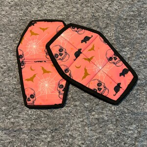 Op de afbeelding: Twee roze en zwarte, kistvormige ovenwanten met een Halloween-thema. De ovenwanten hebben een patroon van schedels, vleermuizen en spinnenwebben.