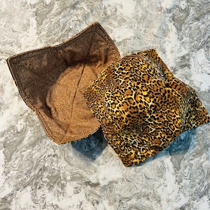 May include: Two fabric bowl cozies. One is brown with a repeating circular pattern. The other is a leopard print in shades of brown, tan, and black. The cozies are square-shaped and designed to hold bowls.