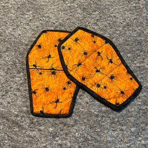 May include: Two orange and black fabric pot holders shaped like coffins with spiderwebs and spiders printed on them.