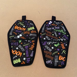 Puede incluir: Dos manoplas de horno con forma de ataúd, de temática de Halloween, en blanco y negro. Las manoplas están cubiertas de una variedad de palabras e imágenes de temática de Halloween, incluyendo "Boo!" "Fun!" "Trick or Treat!" y "Happy Halloween!"
