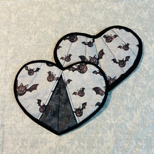 May include: Two heart-shaped Halloween-themed pot holders. The pot holders have a light blue background with a pattern of cartoon bats. They are trimmed with black fabric and have a dark gray lace-like design in the center.