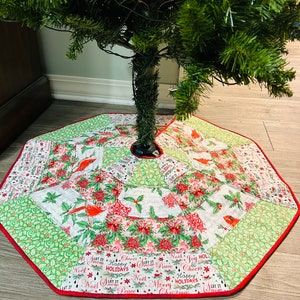 Puede incluir: Una falda de árbol de Navidad verde y blanca con un borde rojo. La falda está decorada con cardenales rojos, acebo y las palabras "Let it Snow", "Happy Holidays", "Merry Christmas", "Peace", "Joy" y "Noel".