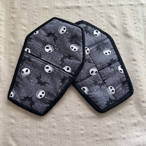 May include: A pair of black and grey oven mitts with a pattern of white Jack Skellington faces and bats. The mitts are shaped like coffins.