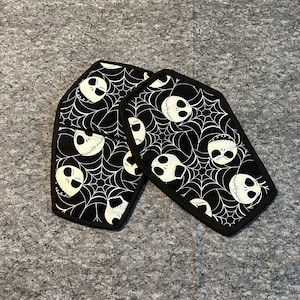 May include: Two black and white pot holders shaped like coffins with a pattern of Jack Skellington's face and spiderwebs.
