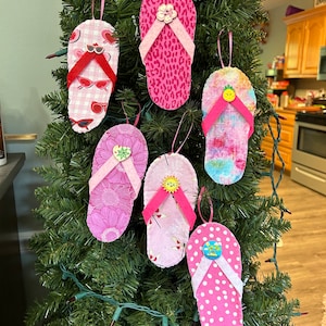 May include: Six pink fabric flip-flop ornaments with different patterns and buttons hanging on a green Christmas tree.